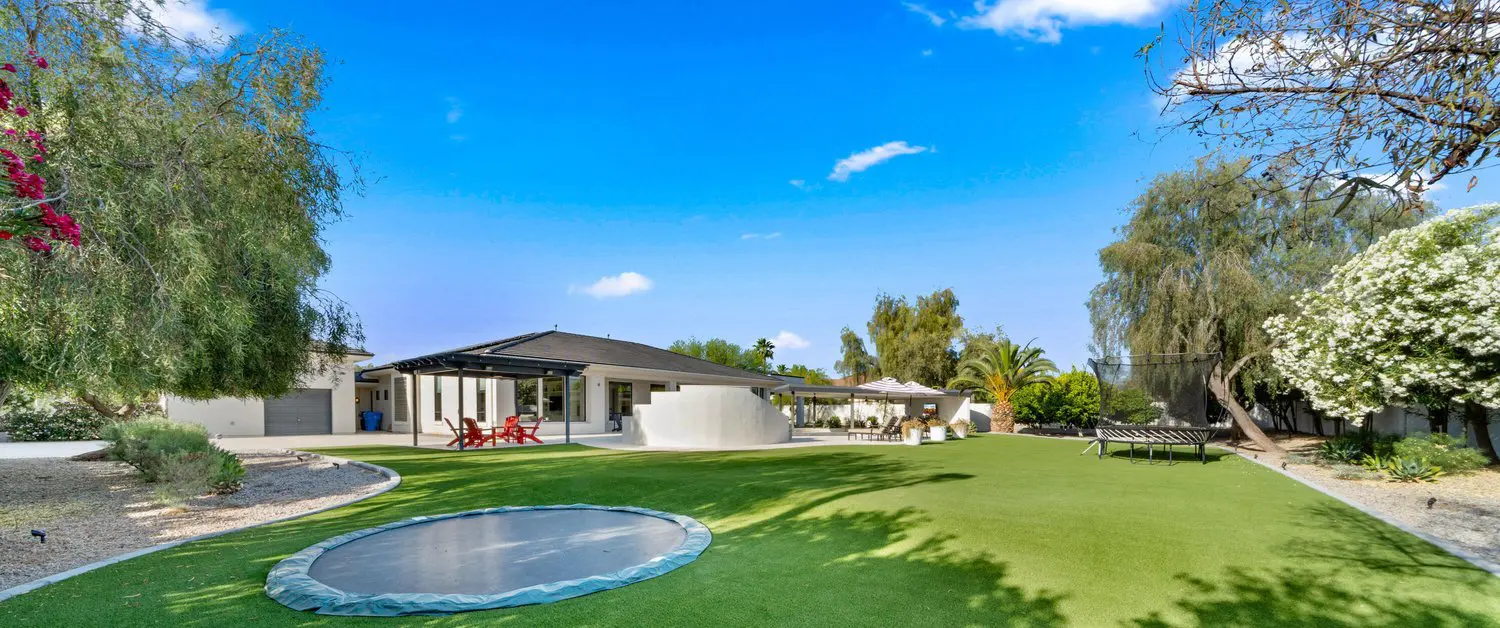 A well-maintained, vibrant green lawn, featuring some areas of synthetic turf, leads up to a two-story house with a beige exterior and brown roof. The house is adorned with various plants and flowers, and a white vehicle is parked to the right. Paver installations border the lush lawn.