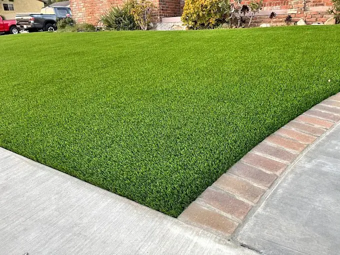 A neatly maintained grass lawn, hinting at the efficiency of artificial turf installation, is bordered by brick edging next to a concrete sidewalk. In Glendale, AZ, a house with brick walls and flourishing plants graces the background.