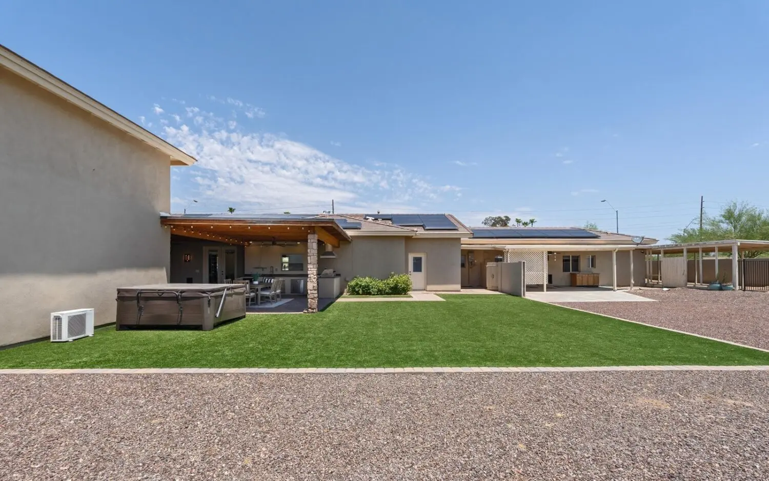 A modern backyard in Glendale, AZ, features a covered patio with outdoor seating, a hot tub, and lush artificial grass. The yard is bordered by a gravel path. A clear blue sky with scattered clouds is visible overhead.