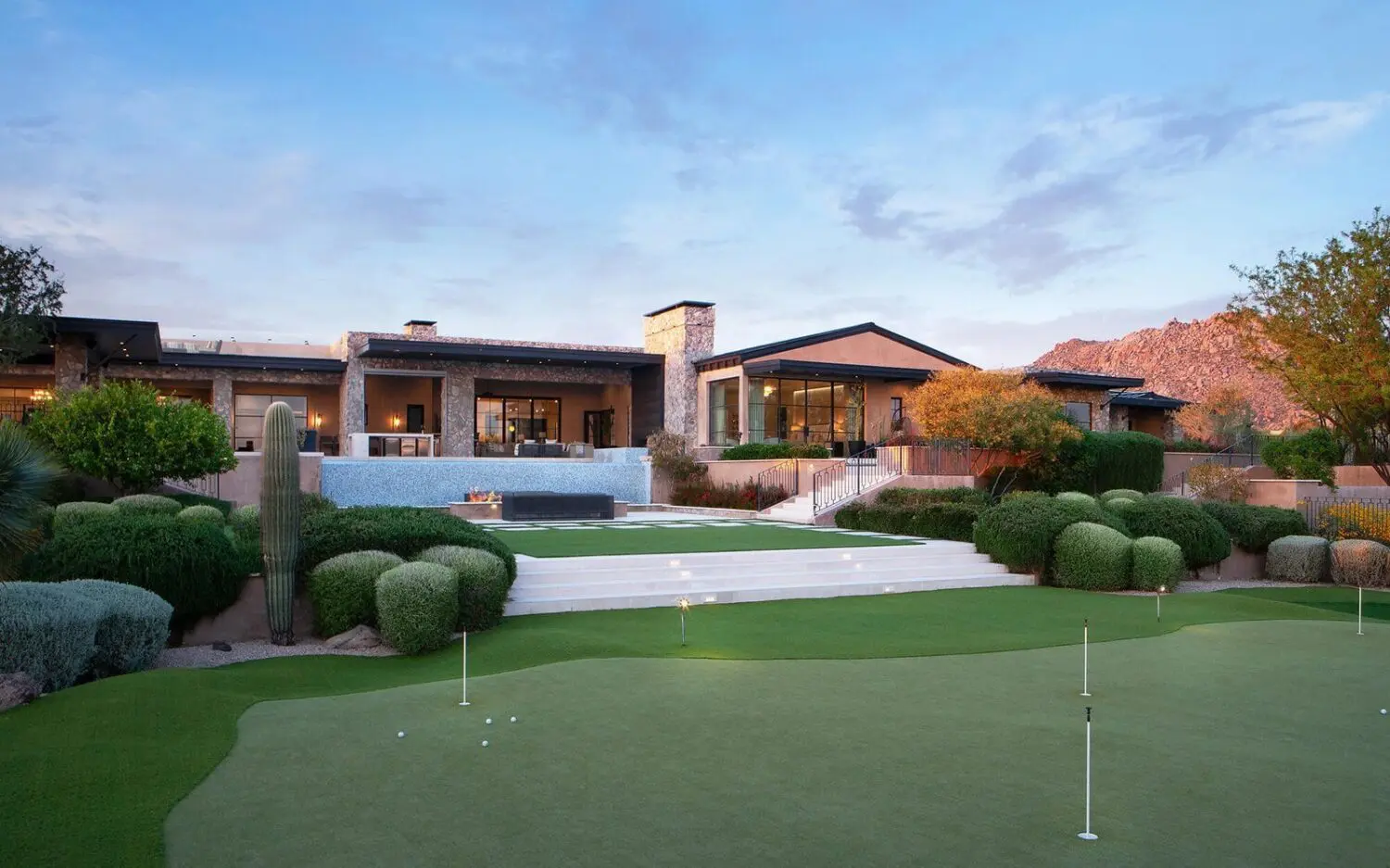 A modern, luxurious house with large windows and multiple sections, set against a backdrop of mountains and clear sky. The foreground boasts a manicured lawn of synthetic turf with a putting green, surrounded by lush greenery and desert plants, perfectly embodying the charm of  Glendale, AZ.