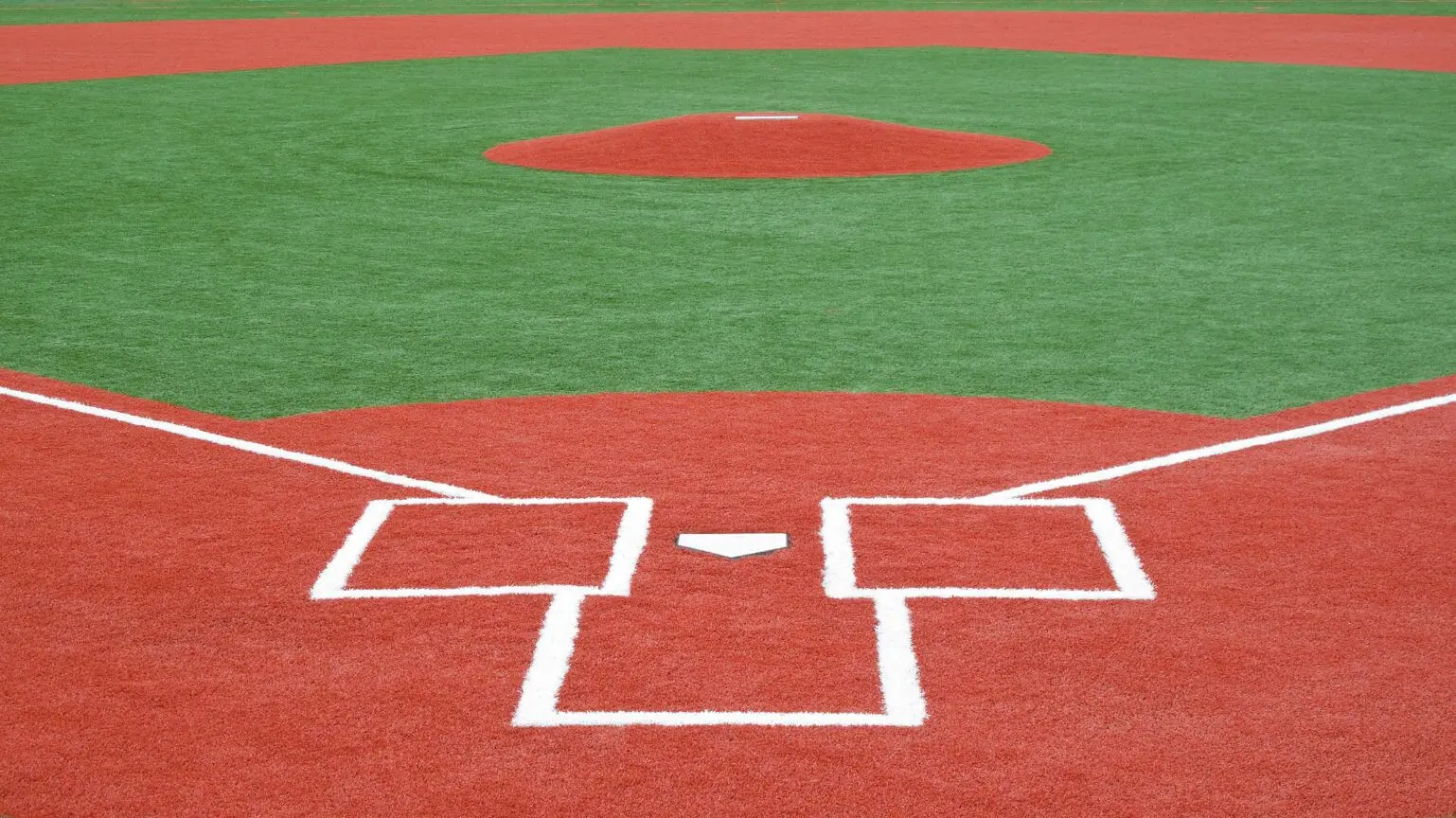 A view of a baseball field from home plate in Sun City, AZ, shows the pitcher's mound at the center. The field boasts synthetic turf solutions with red and green sections, complete with white lines marking the batter's box and bases.