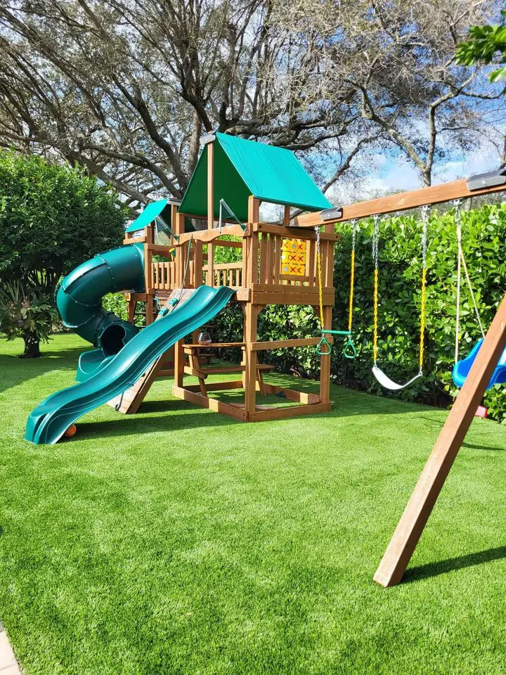 A wooden playset with a green slide, tunnel, and swings rests on a lush lawn enhanced by synthetic turf, surrounded by bushes and trees. The playset features a climbing wall and a peaked roof with canvas under a partly cloudy sky.