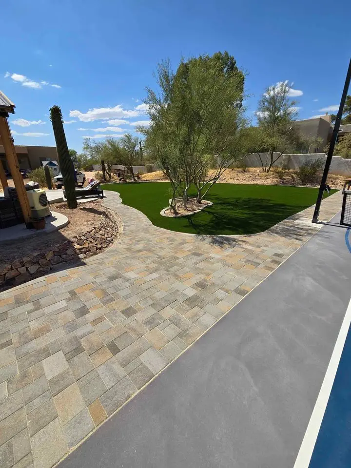 A backyard in Glendale, AZ features a stone pathway leading to a green lawn adorned with desert trees and cacti. With artificial turf installation adding lushness despite the arid climate, a fence and rocky area elegantly border the yard. The clear sky offers partial shade from an unseen structure.