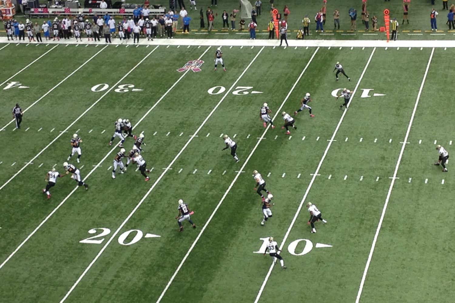 Aerial view of a football game on artificial turf in Glendale, AZ. One team, wearing black, is on offense and advancing the ball, while the other team is in white and defending. The field markings indicate play near the 20-yard line.