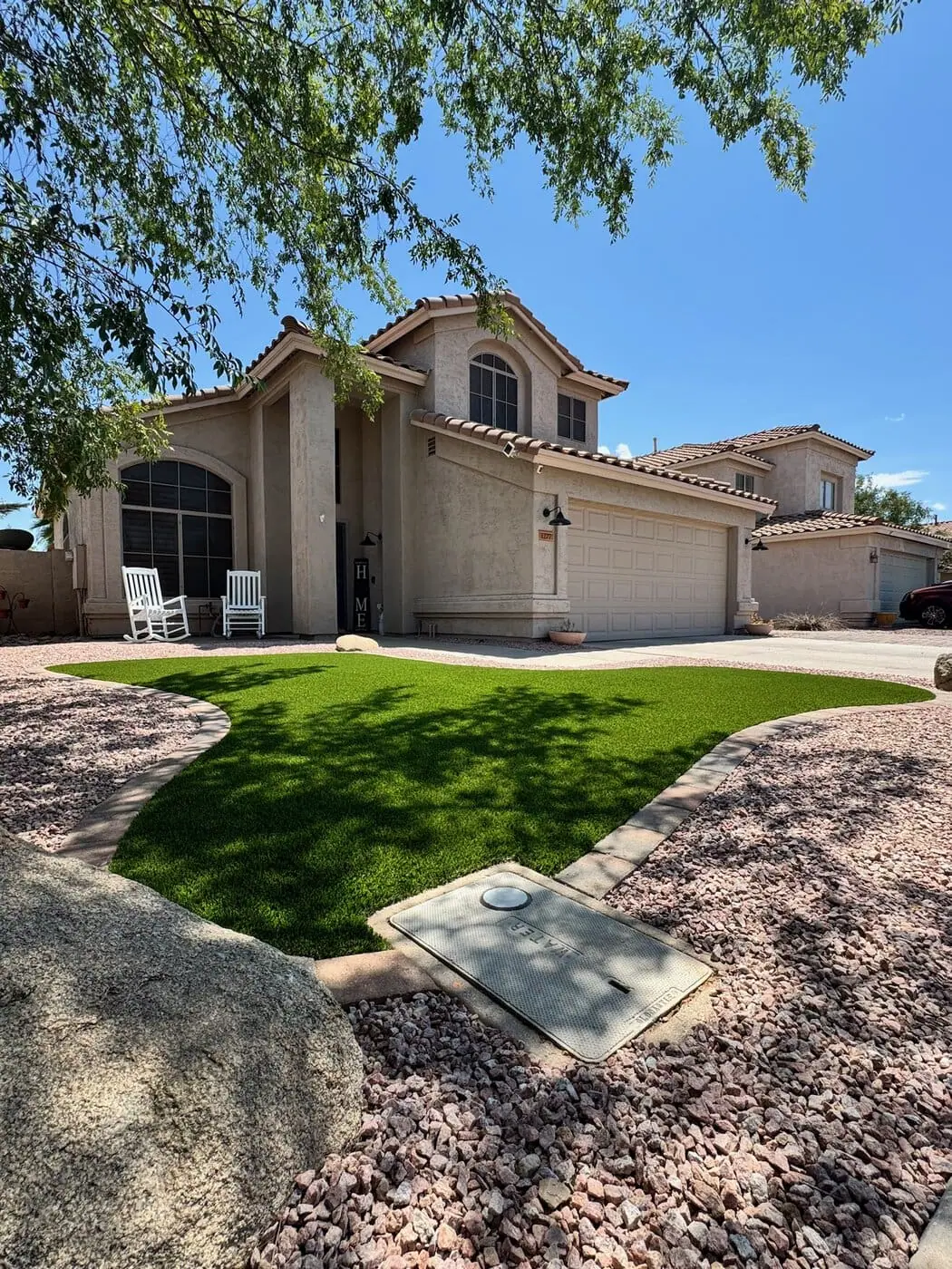 A spacious backyard featuring a green lawn with an in-ground trampoline, shaded seating area with umbrellas, and a modern house with large windows in the background. With synthetic turf installations enhancing its beauty, the yard is surrounded by trees and bushes under a clear blue sky.