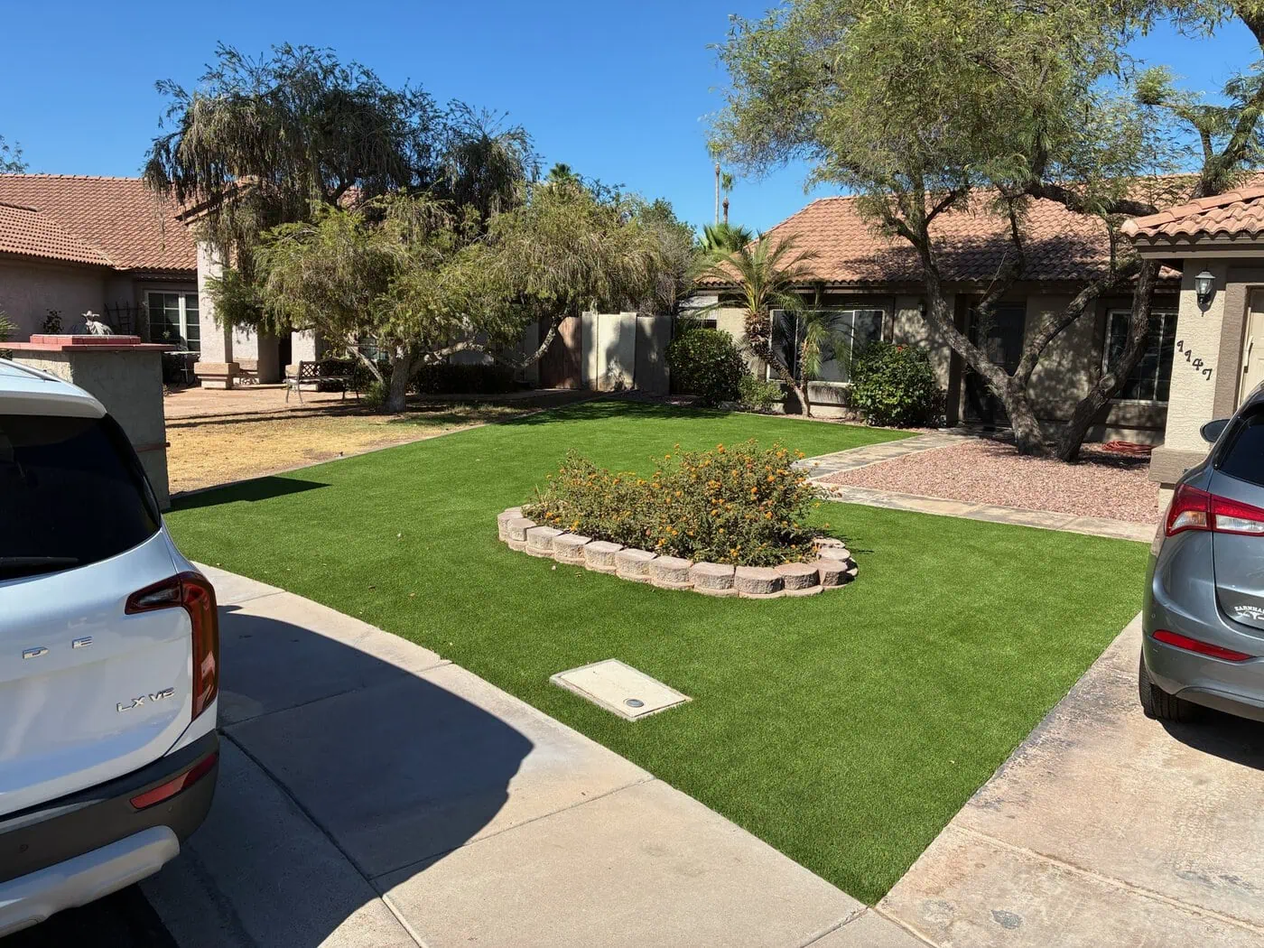 A modern outdoor space featuring grassy areas and sleek paver installations intersected by large square concrete tiles. The courtyard, with its contemporary design, leads to an interior living area visible through floor-to-ceiling glass walls. The space is illuminated by recessed lighting.